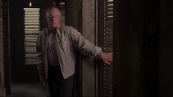 Movie still from “The Way of the Gun” (2000), directed by Christopher McQuarrie – An older man leaning against a wall in front of a window; Medium shot, Low angle