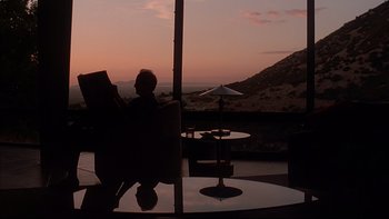 Movie still from “The Way of the Gun” (2000), directed by Christopher McQuarrie – A man sitting in a chair reading a book; Wide shot, Low angle