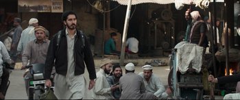 Movie still from “The Wedding Guest” (2018), directed by Michael Winterbottom – A group of men sitting on the ground in front of a building; Wide shot, Over the shoulder angle