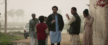 Movie still from “The Wedding Guest” (2018), directed by Michael Winterbottom – A group of men standing on top of a dirt field; Medium shot, Over the shoulder angle