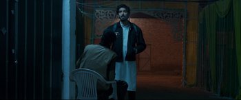 Movie still from “The Wedding Guest” (2018), directed by Michael Winterbottom – A man standing next to a chair in a dark room; Medium shot, Over the shoulder angle