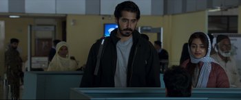 Movie still from “The Wedding Guest” (2018), directed by Michael Winterbottom – A man standing in front of an airport terminal; Medium shot, Over the shoulder angle