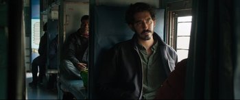 Movie still from “The Wedding Guest” (2018), directed by Michael Winterbottom – A man with a goatee standing on a train; Medium shot, Over the shoulder angle