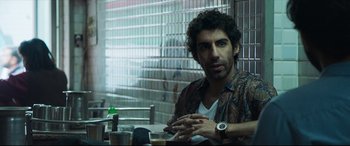 Movie still from “The Wedding Guest” (2018), directed by Michael Winterbottom – A man sitting in front of a window with his hands crossed; Medium shot, Over the shoulder angle