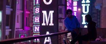 Movie still from “The Wedding Guest” (2018), directed by Michael Winterbottom – A man sitting on a railing in front of a neon sign; Wide shot, Low angle