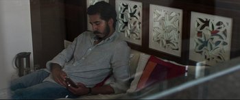 Movie still from “The Wedding Guest” (2018), directed by Michael Winterbottom – A man sitting on top of a couch looking at his cell phone; Medium shot, High angle