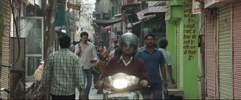Movie still from “The Wedding Guest” (2018), directed by Michael Winterbottom – A group of people walking down a street on a motorcycle; Wide shot, Over the shoulder angle