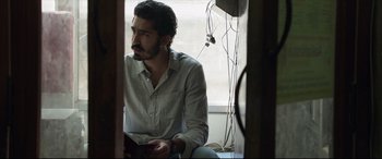 Movie still from “The Wedding Guest” (2018), directed by Michael Winterbottom – A man sitting in front of a window looking at something; Close Up shot, Low angle