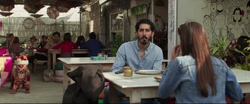 Movie still from “The Wedding Guest” (2018), directed by Michael Winterbottom – A man sitting at an outdoor table with a cup of coffee; Medium shot, Over the shoulder angle