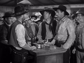 Movie still from “The Westerner” (1940), directed by William Wyler – A black and white photo of a group of men standing around a bar; Medium shot, Over the shoulder angle