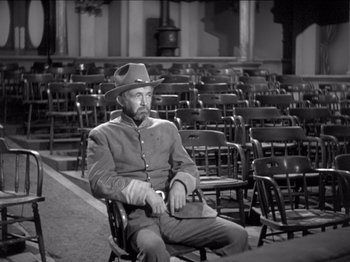 Movie still from “The Westerner” (1940), directed by William Wyler – A man sitting in front of rows of chairs in an auditorium; Medium shot, High angle