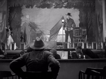 Movie still from “The Westerner” (1940), directed by William Wyler – A man sitting in front of a painting of a man in a cowboy hat; Wide shot, Low angle