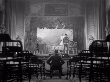 Movie still from “The Westerner” (1940), directed by William Wyler – A man sitting in front of a stage in a theater; Wide shot, High angle