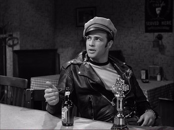 Movie still from “The Wild One” (1953), directed by Laslo Benedek – A black and white photo of a man sitting at a table with a beer; Medium shot, Over the shoulder angle