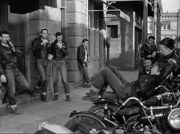 Movie still from “The Wild One” (1953), directed by Laslo Benedek – A black and white photo of a group of men standing around a parked motorcycle; Wide shot, High angle