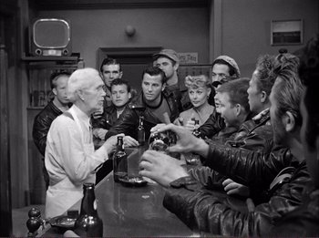 Movie still from “The Wild One” (1953), directed by Laslo Benedek – An old man is sitting at a bar with a group of men; Medium shot, High angle