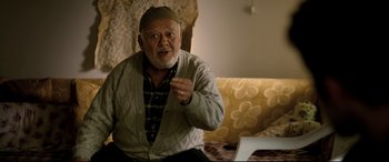 Movie still from “The Wild Pear Tree” (2018), directed by Nuri Bilge Ceylan – An older man sitting on a couch in front of a computer; Medium shot, Over the shoulder angle
