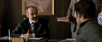 Movie still from “The Wild Pear Tree” (2018), directed by Nuri Bilge Ceylan – A man sitting at a desk in front of another man holding a book; Medium shot, Over the shoulder angle