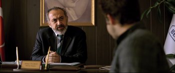Movie still from “The Wild Pear Tree” (2018), directed by Nuri Bilge Ceylan – A man sitting at a table talking to another man in front of him; Medium shot, Over the shoulder angle