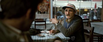 Movie still from “The Wild Pear Tree” (2018), directed by Nuri Bilge Ceylan – An older man sitting at a table in front of a glass of water; Medium shot, Over the shoulder angle