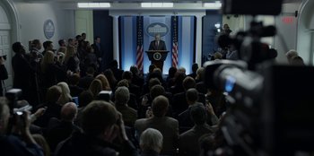 Movie still from “House of Cards” (2013), created by Beau Willimon – A crowd of people watching a man in a suit speaking at a podium; Wide shot, Over the shoulder angle