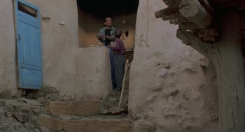 Movie still from “The Wind Will Carry Us” (1999), directed by Abbas Kiarostami – A man and a woman are standing in a doorway; Wide shot, Low angle