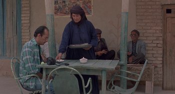 Movie still from “The Wind Will Carry Us” (1999), directed by Abbas Kiarostami – An old woman is serving food at an outdoor table; Wide shot, Over the shoulder angle