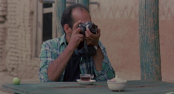 Movie still from “The Wind Will Carry Us” (1999), directed by Abbas Kiarostami – A man sitting at a table taking a picture with a camera; Close Up shot, Over the shoulder angle
