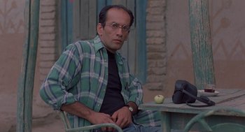 Movie still from “The Wind Will Carry Us” (1999), directed by Abbas Kiarostami – A man sitting at a table with an apple on it; Medium shot, Low angle