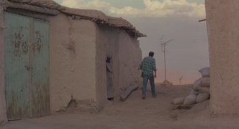 Movie still from “The Wind Will Carry Us” (1999), directed by Abbas Kiarostami – A man standing in front of an adobe building; Wide shot, Low angle