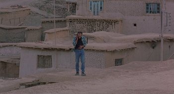 Movie still from “The Wind Will Carry Us” (1999), directed by Abbas Kiarostami – A man standing on a dirt road in front of a building; Wide shot, High angle