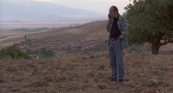 Movie still from “The Wind Will Carry Us” (1999), directed by Abbas Kiarostami – A man standing on a rocky hillside talking on a cell phone; Wide shot, Low angle