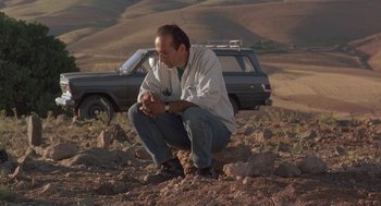 Movie still from “The Wind Will Carry Us” (1999), directed by Abbas Kiarostami – A man crouching on the ground in front of a car; Wide shot, Low angle