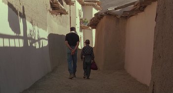 Movie still from “The Wind Will Carry Us” (1999), directed by Abbas Kiarostami – A man and a child walking down a street; Wide shot, Low angle