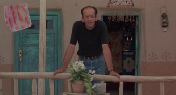 Movie still from “The Wind Will Carry Us” (1999), directed by Abbas Kiarostami – A man leaning on a railing with a potted plant on top; Medium shot, Low angle