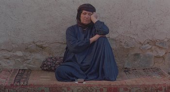 Movie still from “The Wind Will Carry Us” (1999), directed by Abbas Kiarostami – An older woman sitting on the ground next to a cup; Medium shot, High angle