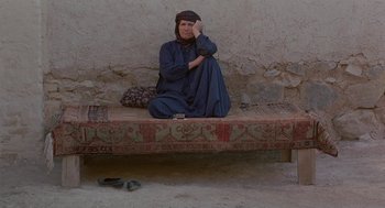 Movie still from “The Wind Will Carry Us” (1999), directed by Abbas Kiarostami – An older woman sitting on a bed in a village; Wide shot, High angle