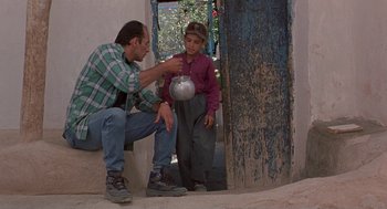 Movie still from “The Wind Will Carry Us” (1999), directed by Abbas Kiarostami – A man sitting on a wall next to a young boy; Wide shot, Low angle
