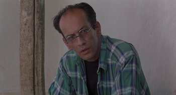 Movie still from “The Wind Will Carry Us” (1999), directed by Abbas Kiarostami – A person wearing glasses and a plaid shirt; Close Up shot, Low angle