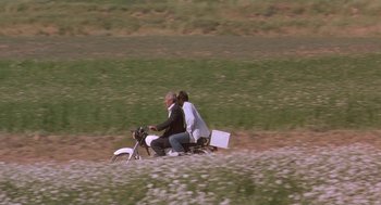 Movie still from “The Wind Will Carry Us” (1999), directed by Abbas Kiarostami – A man and a woman riding a motorcycle through a field; Wide shot, High angle