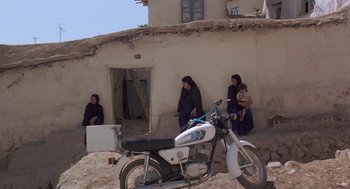 Movie still from “The Wind Will Carry Us” (1999), directed by Abbas Kiarostami – A motorcycle parked in front of a building with people sitting on it; Wide shot, High angle