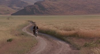 Movie still from “The Wind Will Carry Us” (1999), directed by Abbas Kiarostami – Two people riding a motorcycle down a dirt road; Extreme Wide shot, High angle