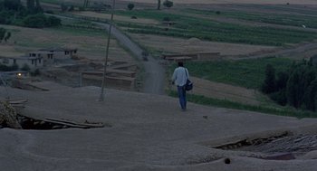 Movie still from “The Wind Will Carry Us” (1999), directed by Abbas Kiarostami – A man walking down a dirt road in a field; Extreme Wide shot, High angle
