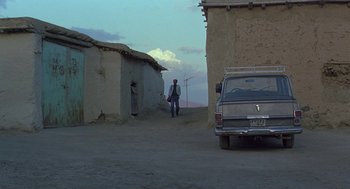 Movie still from “The Wind Will Carry Us” (1999), directed by Abbas Kiarostami – A man walking down a dirt road near a building; Wide shot, Low angle