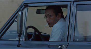 Movie still from “The Wind Will Carry Us” (1999), directed by Abbas Kiarostami – A man sitting in the driver's seat of a car smoking a cigarette; Close Up shot, Low angle