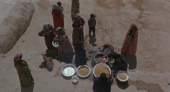 Movie still from “The Wind Will Carry Us” (1999), directed by Abbas Kiarostami – A group of people standing around a pile of food; Extreme Wide shot, Overhead angle