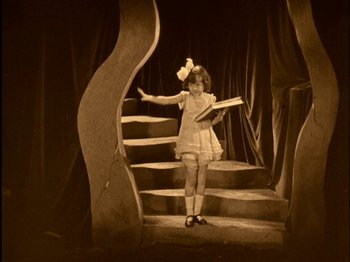 Movie still from “The Wizard of Oz” (1925), directed by Larry Semon – A little girl standing on some steps holding a book; Wide shot, High angle