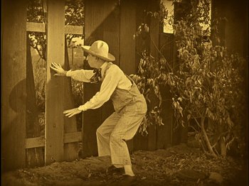 Movie still from “The Wizard of Oz” (1925), directed by Larry Semon – An old photo of a man in a hat and overalls leaning against a fence; Wide shot, High angle