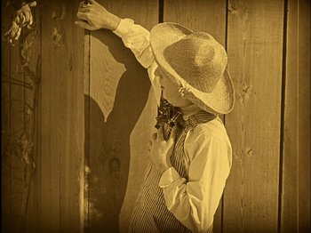 Movie still from “The Wizard of Oz” (1925), directed by Larry Semon – A young man wearing a straw hat and a neck tie leaning against a wooden wall; Medium shot, High angle