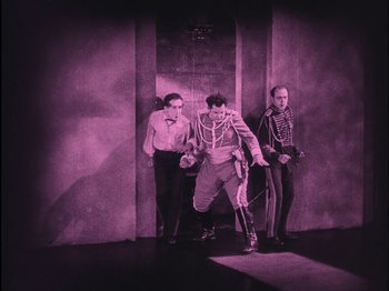 Movie still from “The Wizard of Oz” (1925), directed by Larry Semon – A man in military garb is standing next to two other men; Wide shot, High angle